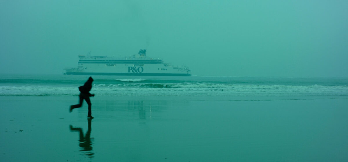 Un migrant courant sur la plage de Calais devant un bateau dans L’Héroïque lande