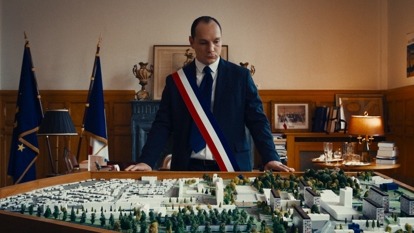 Le maire (Alexis Manenti) regarde la maquette des constructions dans Bâtiment 5